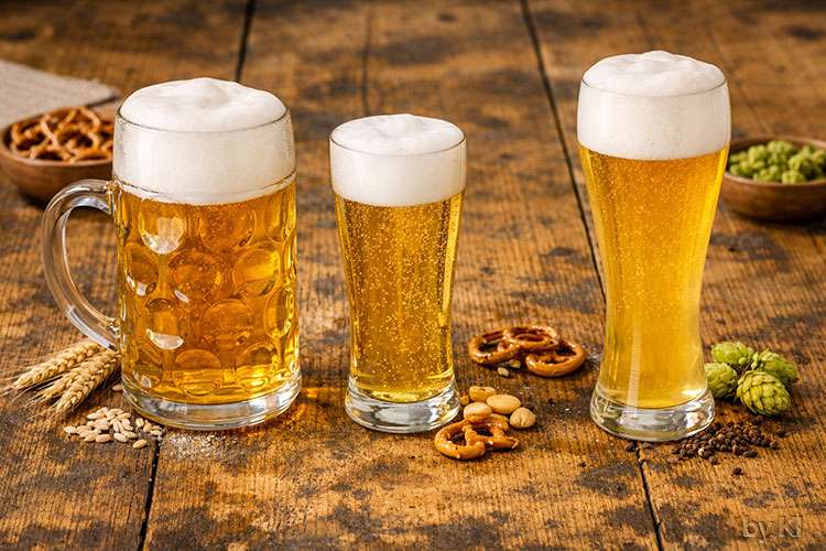Drei Biere stehen nebeneinander auf einem rustikalen Holztisch:</p>
<p>Maß Bier: Ein großer Maßkrug mit Henkel, gefüllt mit goldgelbem Bier und einer kräftigen, weißen Schaumkrone. Das Glas ist dickwandig und wirkt schwer, das Bier klar und spritzig.</p>
<p>Pils: Ein schlankes, hohes Glas mit hellgoldenem Bier, feinperlig und mit einer kompakten, stabilen Schaumhaube. Der Duft ist frisch und leicht herb.</p>
<p>Weizenbier: Ein hohes, geschwungenes Glas mit trüb‑goldenem Bier, die Schaumkrone ist besonders luftig und cremig. Der Eindruck ist mild, fruchtig und weich.</p>
<p>Das Licht ist warm und natürlich, die Stimmung einladend und authentisch – typisch für eine gemütliche Bierverkostung oder Wirtshaus‑Szene.