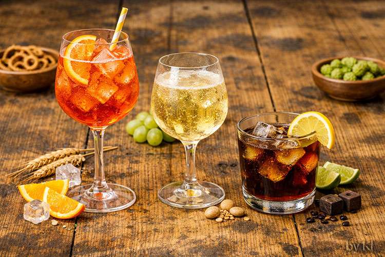 Drei Longdrinks stehen nebeneinander auf einem rustikalen Holztisch:</p>
<p>Aperol Spritz: Ein großes Weinglas mit leuchtend orangefarbenem Getränk, gefüllt mit Eiswürfeln und einer Orangenscheibe am Glasrand. Das Getränk wirkt spritzig und sommerlich.</p>
<p>Weinschorle: Ein bauchiges Glas mit hellgoldenem, leicht perlendem Getränk, feine Bläschen steigen nach oben. Der Eindruck ist frisch und leicht.</p>
<p>Whisky Cola: Ein kurzes, breites Glas mit dunkelbraunem Getränk, Eiswürfeln und einer Zitronenscheibe. Der Duft ist süßlich und kräftig.</p>
<p>Das Licht ist warm und natürlich, die Stimmung entspannt und sommerlich, ideal für eine Getränkekarte oder Bar‑Szene.