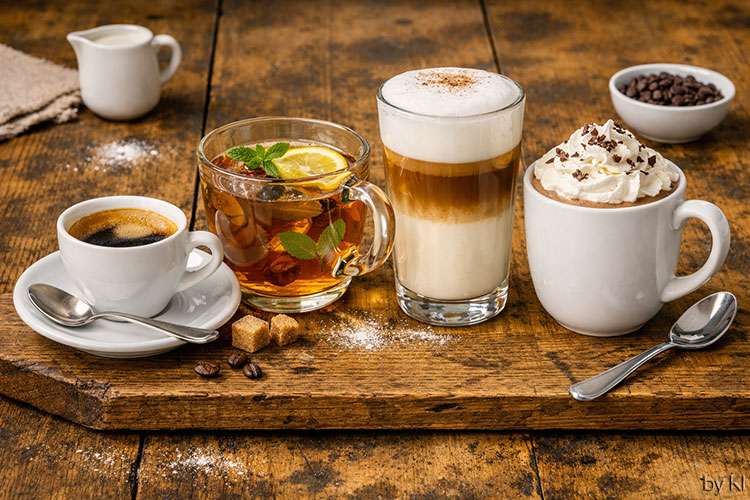 Heiße Getränke stehen nebeneinander auf einem rustikalen Holztisch:<br />
Kaffee: Dunkelbraun mit feiner, goldener Crema in einer weißen Tasse. Der Duft ist kräftig und aromatisch.<br />
Tee: Goldfarben und klar, mit einer Zitronenscheibe und frischen Minzblättern. Der Geruch ist frisch und leicht fruchtig.<br />
Latte Macchiato: Im hohen Glas geschichtet – unten Milch, in der Mitte Espresso, oben cremiger Milchschaum mit einem Hauch Kaffeepulver.<br />
Heiße Schokolade: Dickflüssig und samtig, mit einer Sahnehaube und Schokoraspeln. Der Duft ist süß und warm.<br />
Im Hintergrund stehen kleine Schälchen mit Milch, Kaffeebohnen und Zuckerwürfeln. Das Licht ist hell und natürlich, die Stimmung einladend und gemütlich.