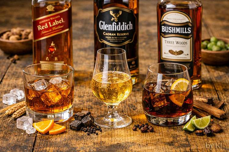Drei Whiskys stehen nebeneinander auf einem rustikalen Holztisch, jeder in einem passenden Glas mit eigener Farbe und Charakter:</p>
<p>Johnnie Walker Red Label: Ein klassischer Tumbler mit dickem Boden, gefüllt mit goldenem Whisky und Eiswürfeln. Das Getränk wirkt klar und lebendig, mit leichtem Rauchduft und würziger Note.</p>
<p>Glenfiddich Caoran Reserve: Ein Glencairn‑Glas mit goldgelbem Whisky, leicht ölig und aromatisch. Der Duft ist warm, mit Anklängen von Torf und Malz.</p>
<p>Bushmills Three Woods: Ein rundes Snifter‑Glas mit tief bernsteinfarbenem Whisky, leicht viskos, mit Noten von Holz und Trockenfrüchten.</p>
<p>Im Hintergrund stehen die dazugehörigen Flaschen, deren Etiketten farblich abgestimmt sind: Rot, Schwarz und Creme. Auf dem Tisch liegen Eiswürfel, Orangenscheiben, Hopfen, Holzstücke und Rosinen als Dekoration.</p>
<p>Das Licht ist warm und natürlich, die Stimmung ruhig, edel und authentisch – ideal für eine Whisky‑Verkostung oder eine hochwertige Getränkekarte.